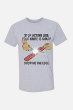White t-shirt featuring a graphic of hands sharpening a knife on a stone with spark effects, with the text “Stop Acting Like Your Knife Is Sharp” above and “Show Me the Edge” below.