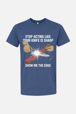 Blue t-shirt featuring a graphic of hands sharpening a knife on a stone with spark effects, with the text “Stop Acting Like Your Knife Is Sharp” above and “Show Me the Edge” below.